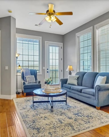 Bright living room with blue sofa, hardwood floors, fireplace, and floor-to-ceiling windows with shutters overlooking water.