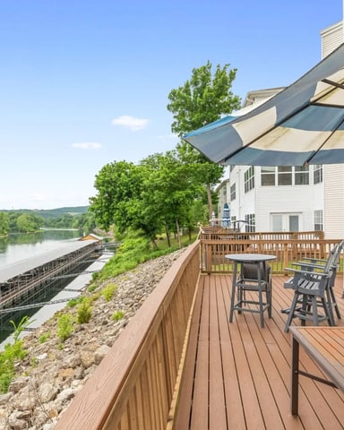 Waterfront deck with striped umbrella, seating, and river view overlooking wooded hillside
