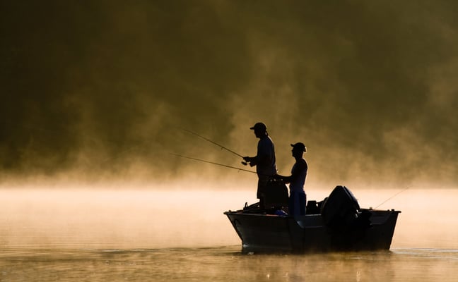 Two people fishing from a small boat on misty water at dawn or dusk